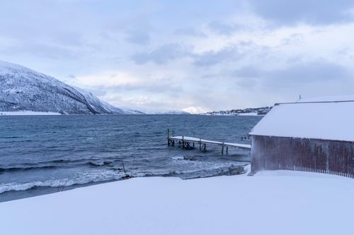 Free A serene arctic scene featuring a snowy coastline with a wooden pier, mountain, and hut in Norway. Stock Photo