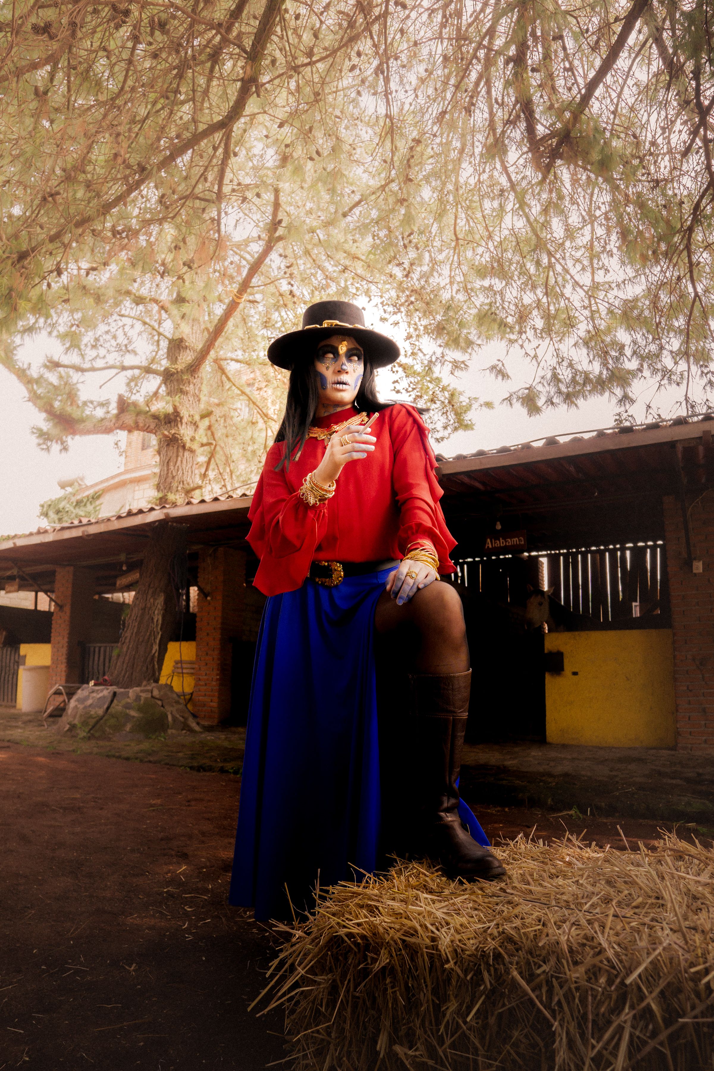 A fashionable woman in a red blouse and blue skirt poses confidently in an outdoor rustic setting.