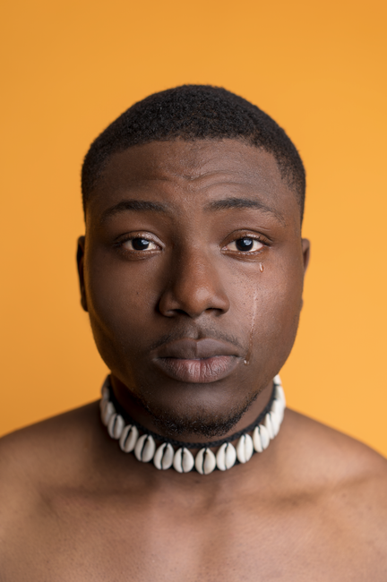 A serious man with a shell necklace sheds a single tear against an orange background.