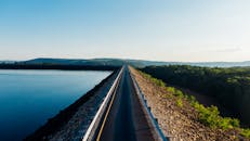 在湛蓝的天空下，森林覆盖的山丘环绕着风景秀丽的湖边高速公路，鸟瞰图景致宜人