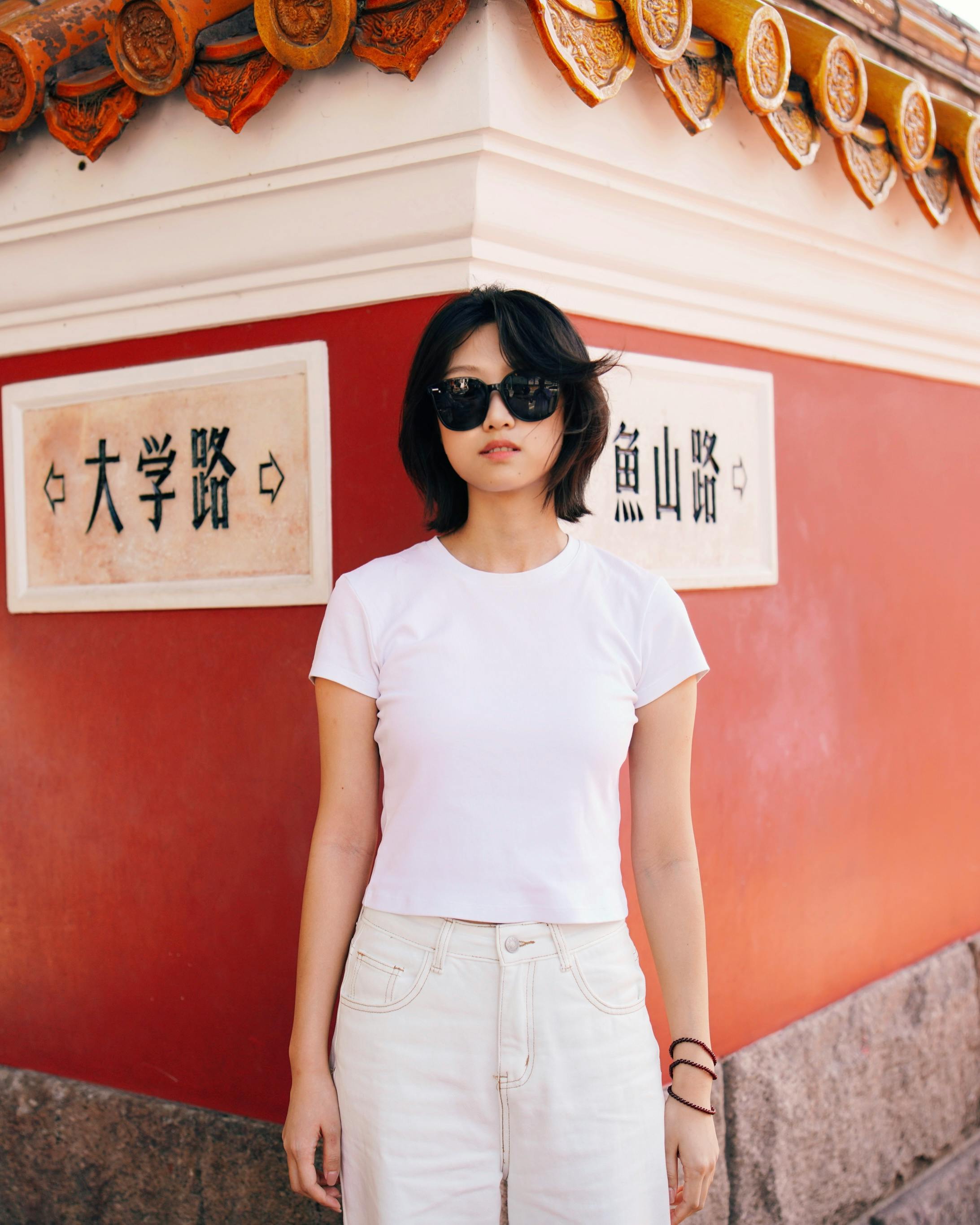 Young woman in sunglasses stands by an Asian-style wall with signage.