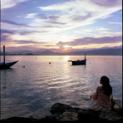 Free A serene scene of a woman watching the sunset by a calm lake with fishing boats. Stock Photo