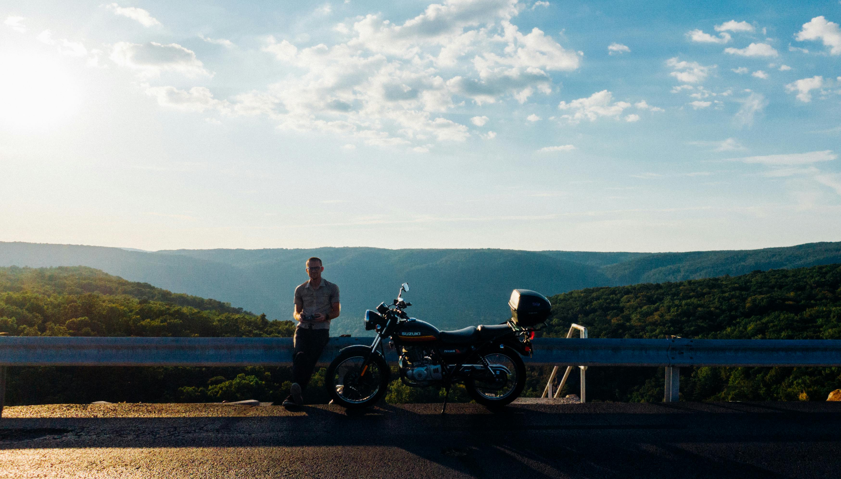 A motorcyclist rests by their bike on a mountain road, enjoying a sunny day in nature.