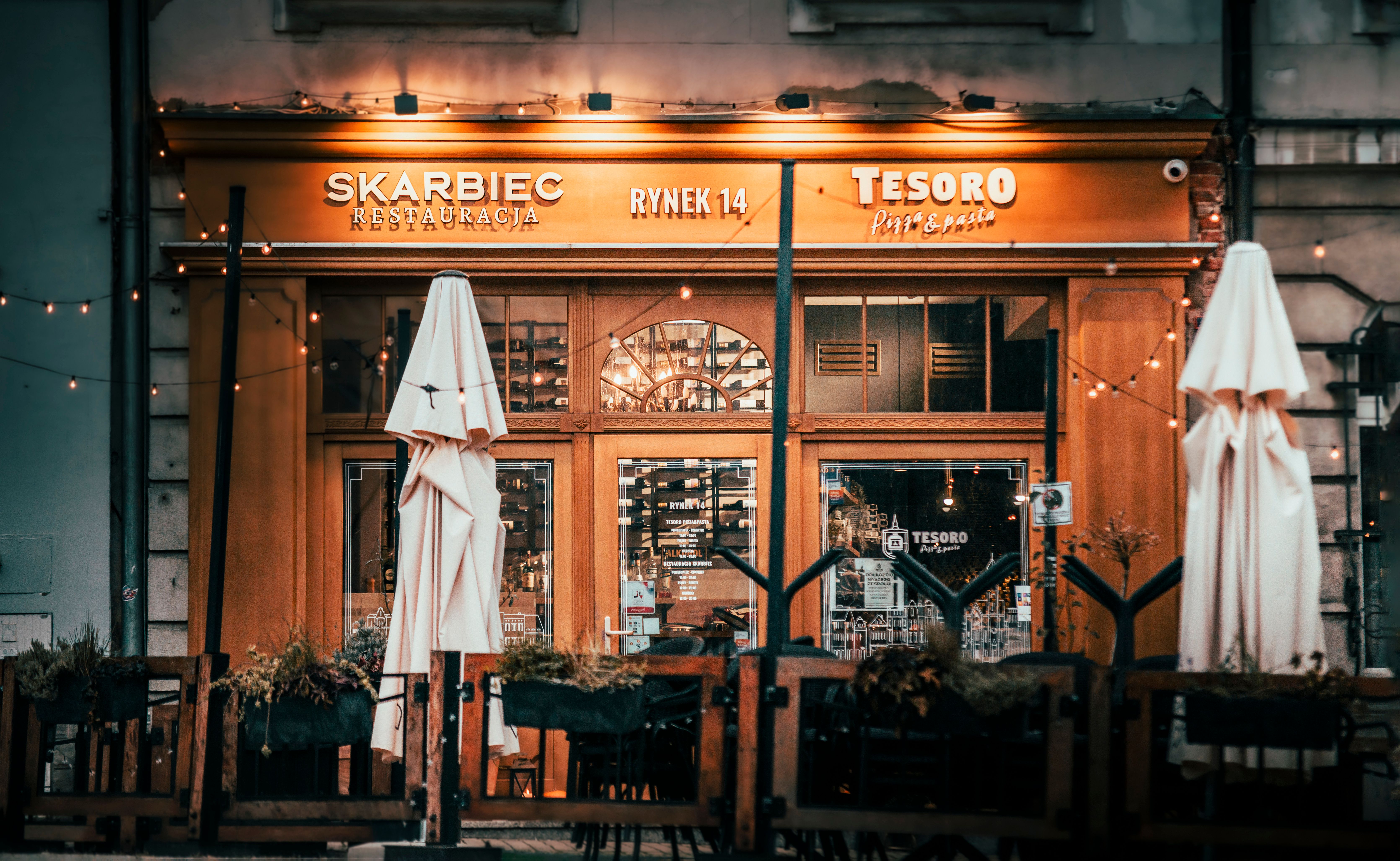 Ciepłe, zachęcające wejście do restauracji Skarbiec z miejscami do siedzenia na zewnątrz i delikatnym oświetleniem, symbolizujące udany biznes i dbałość o detale.