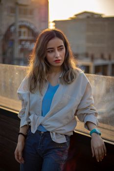 A young woman in casual attire leaning on a railing in a city at sunset.