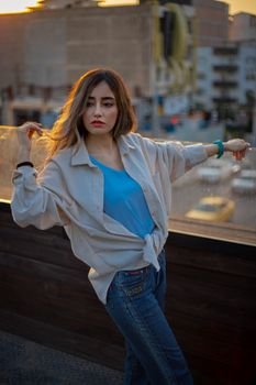 Stylish woman poses casually on urban rooftop, sunset in background.