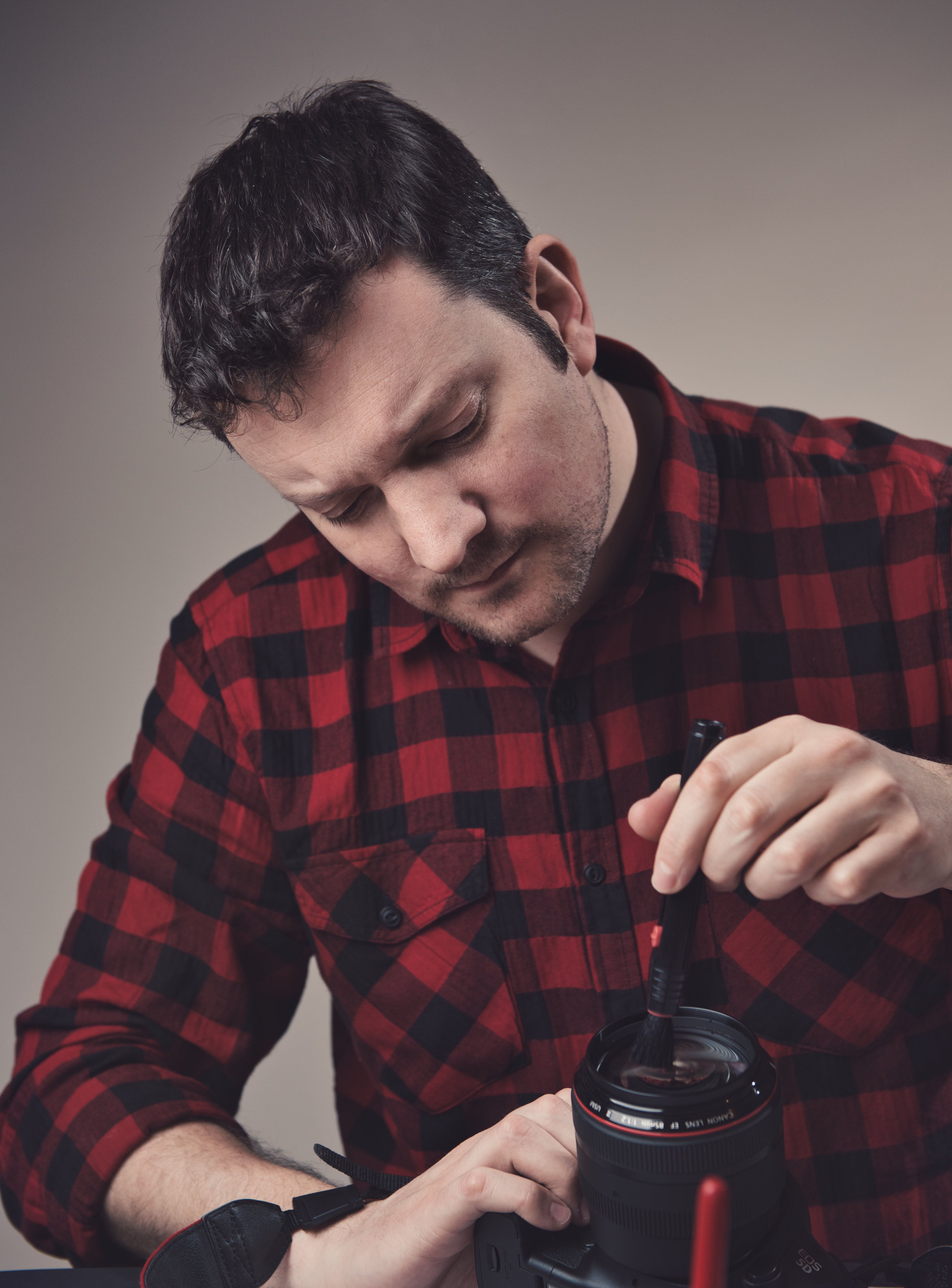 A man in a plaid shirt carefully cleans his DSLR camera lens indoors.