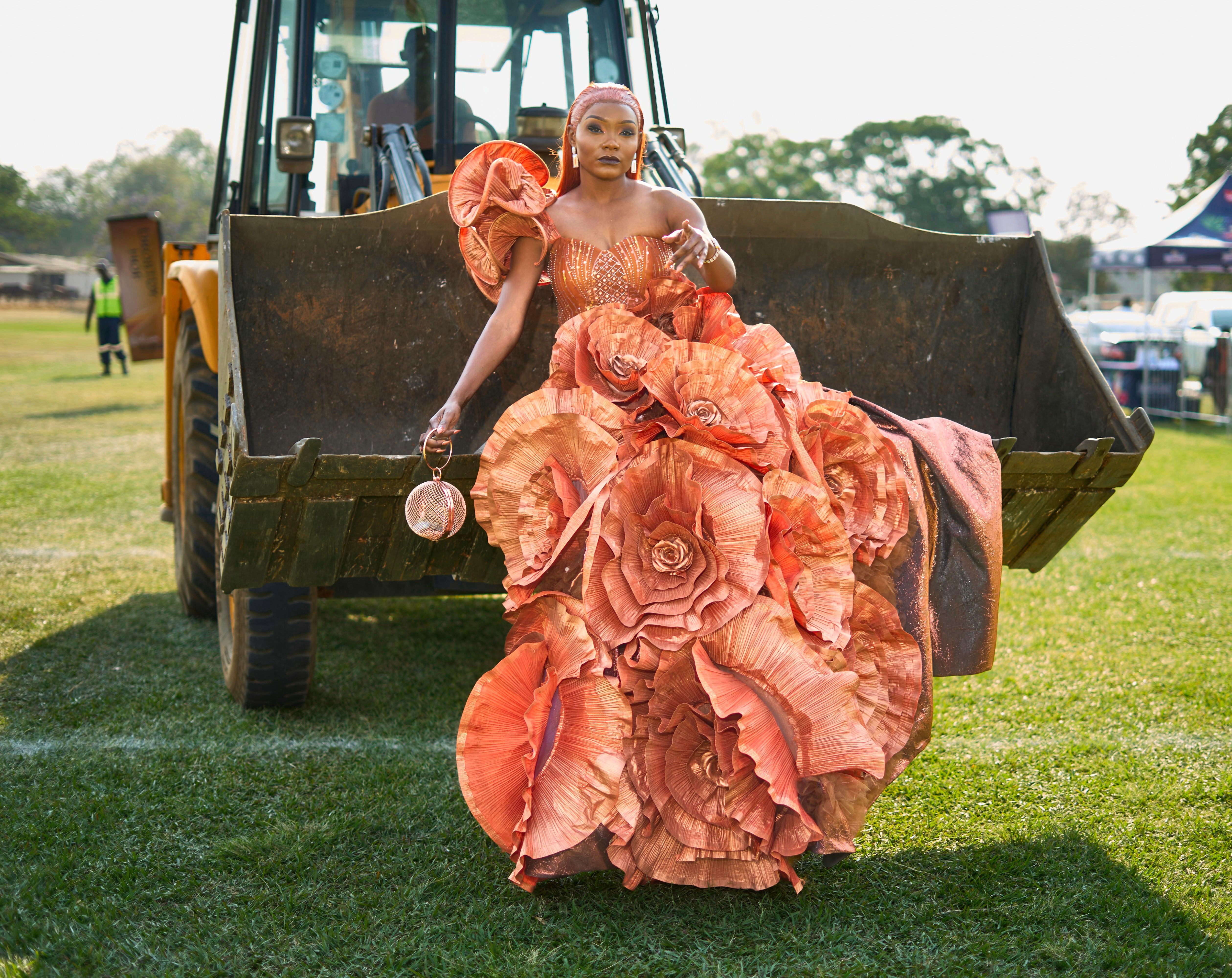 A fashionable woman in a stunning floral dress poses near an excavator on a sunny day in Lusaka.