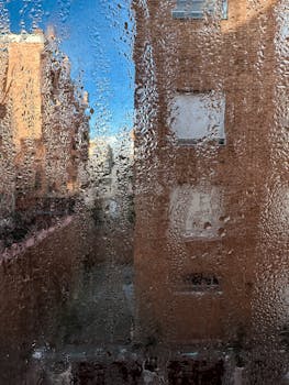 View through a rain-covered window showing urban buildings and clear blue sky in the background.