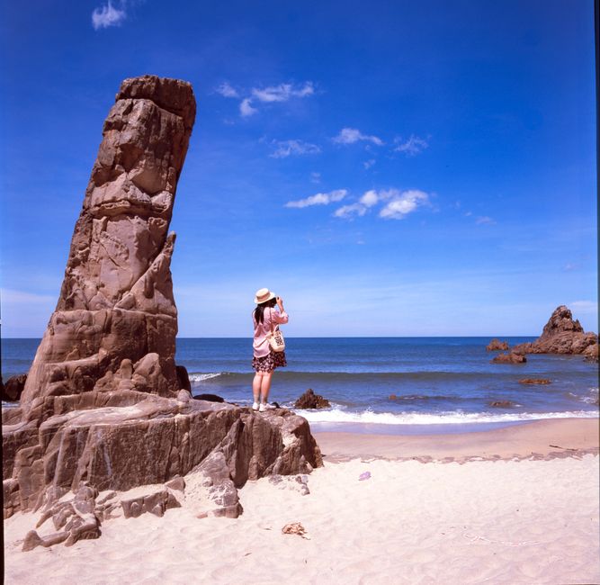 A woman stands on a rock capturing the serene beach view on a sunny day.