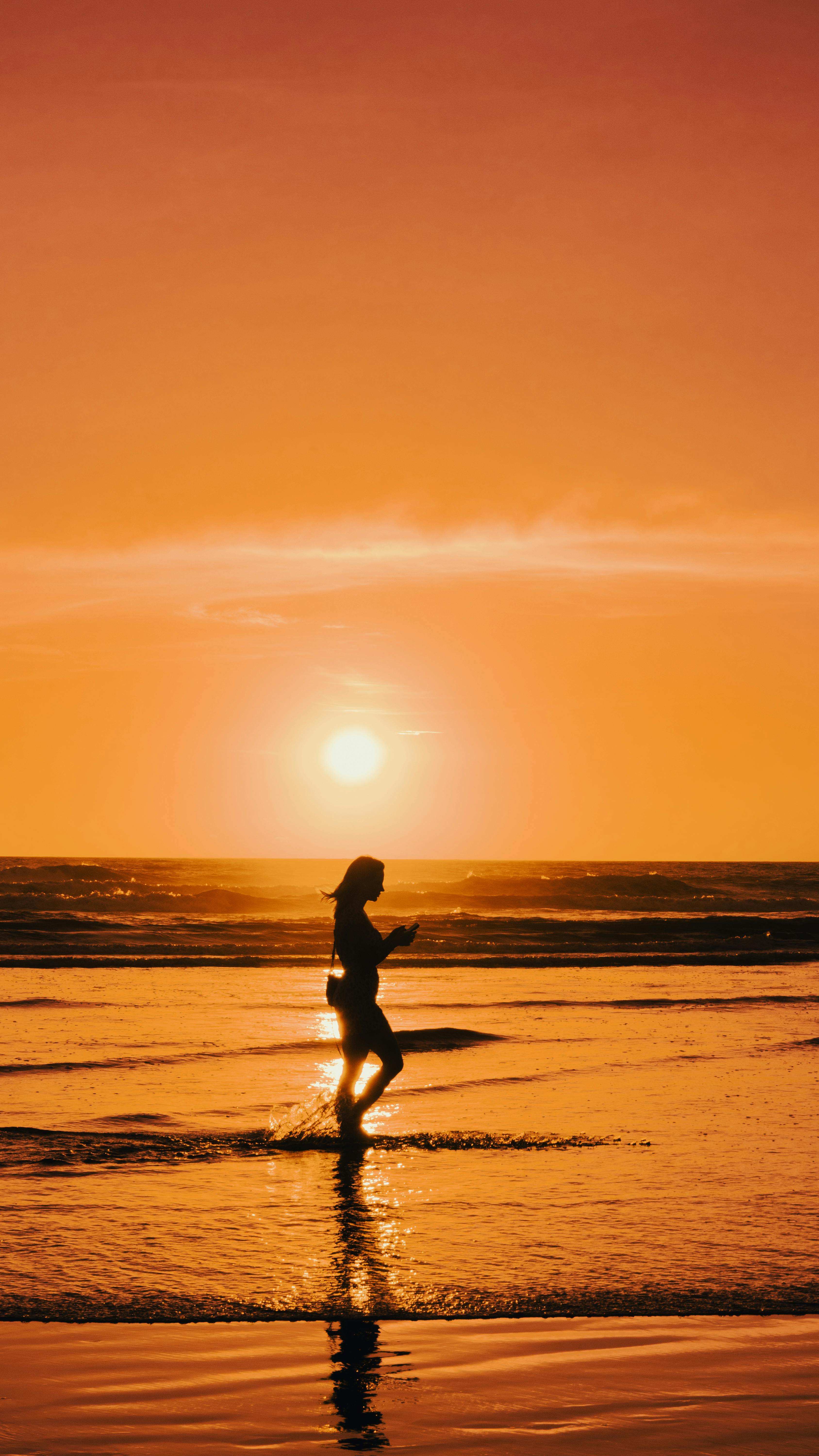 A person runs along the beach during a vibrant sunset, creating a serene silhouette.