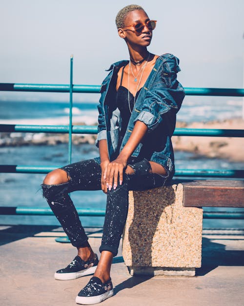 Photo of Woman in Blue Denim Jacket, Black Jeans, and Sunglasses Sitting on Bench Posing While Looking Away