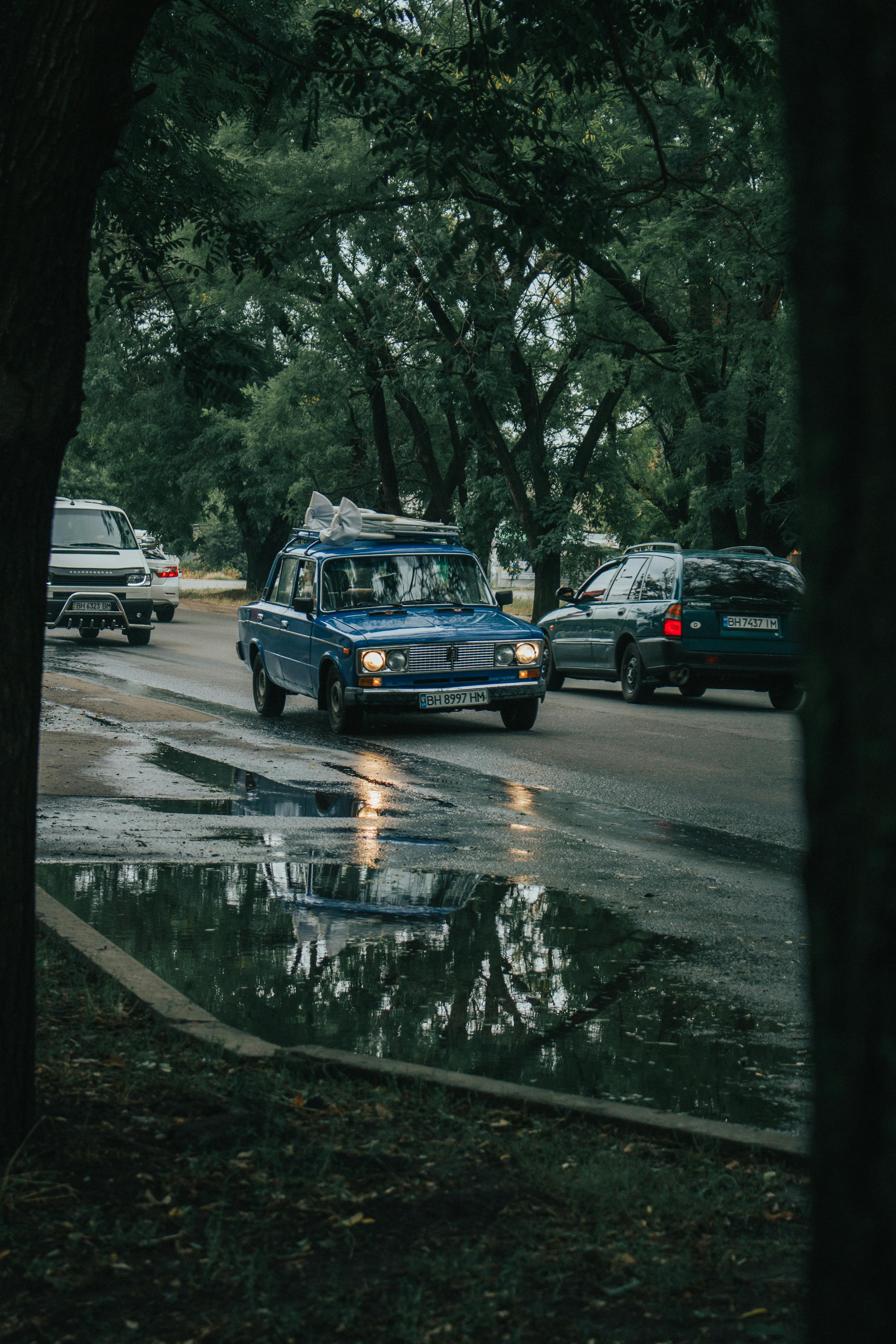 一辆在雨天行驶的老爷车，倒映在被郁郁葱葱的树木包围的水坑里