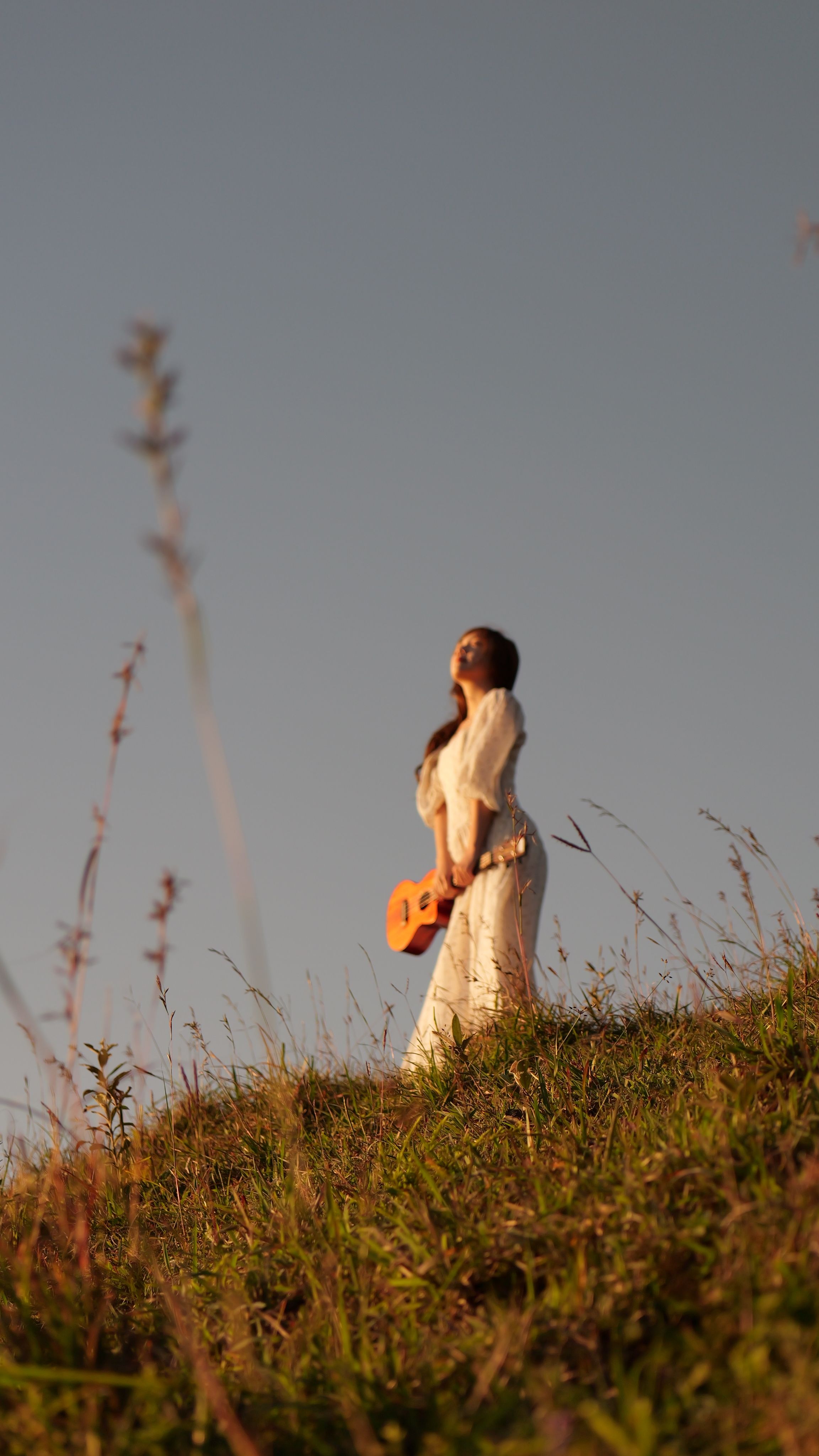 A woman in a white dress stands on a grassy hillside holding a guitar during sunset.