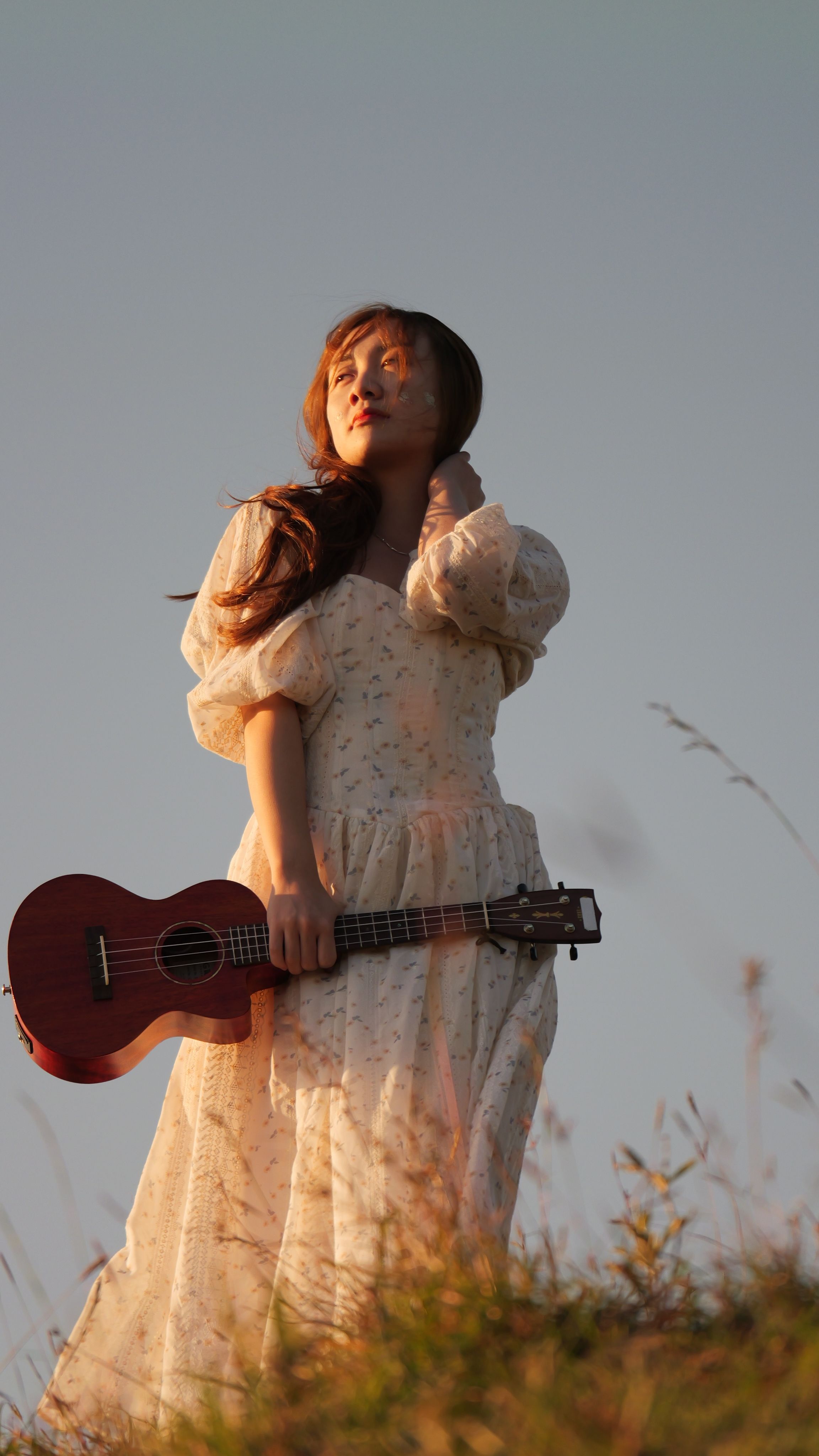 A woman enjoying the outdoors holding a ukulele, dressed in a vintage style gown during sunset.