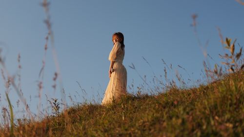 Free A serene scene of a woman in a white dress standing on a grassy hill at sunset. Stock Photo