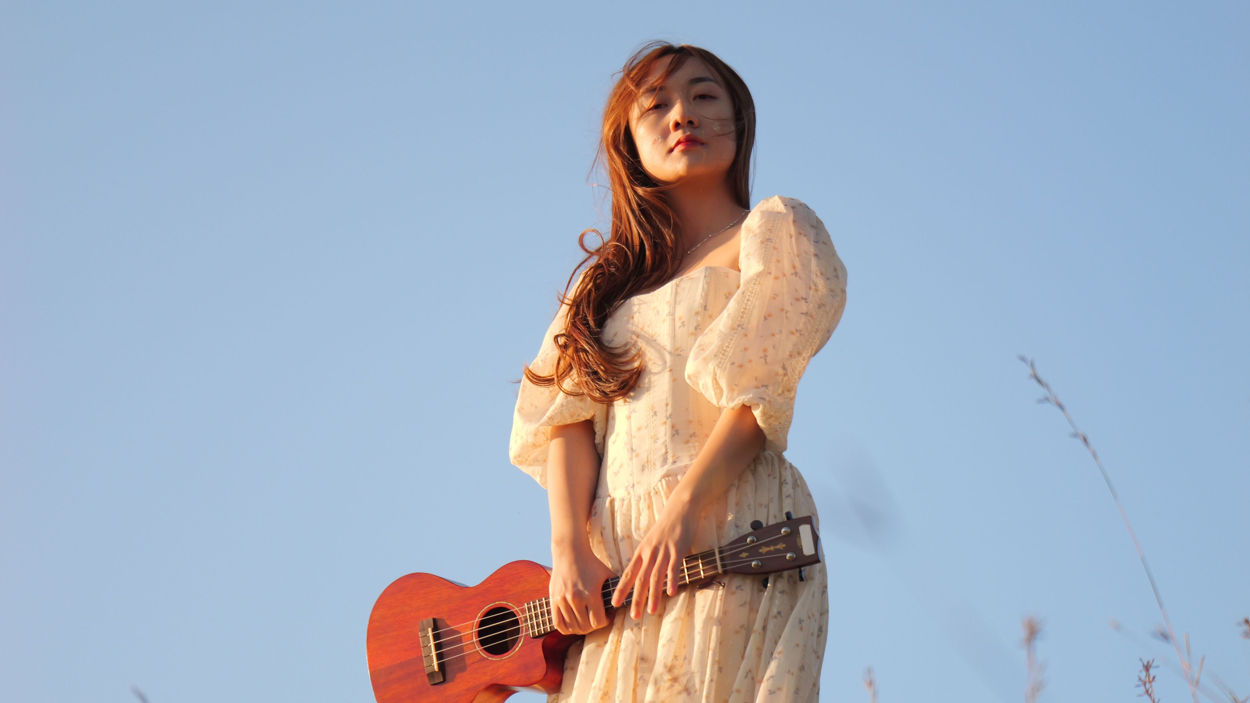 A woman standing outdoors holding a ukulele, gazing thoughtfully, with a clear blue sky backdrop.