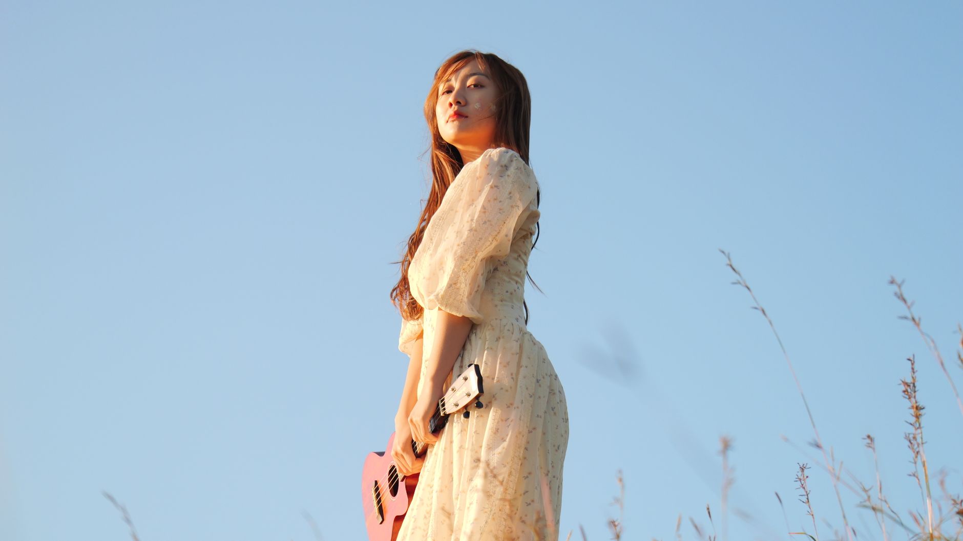 Young woman in a light dress holds a pink ukulele in a sunny, open field.