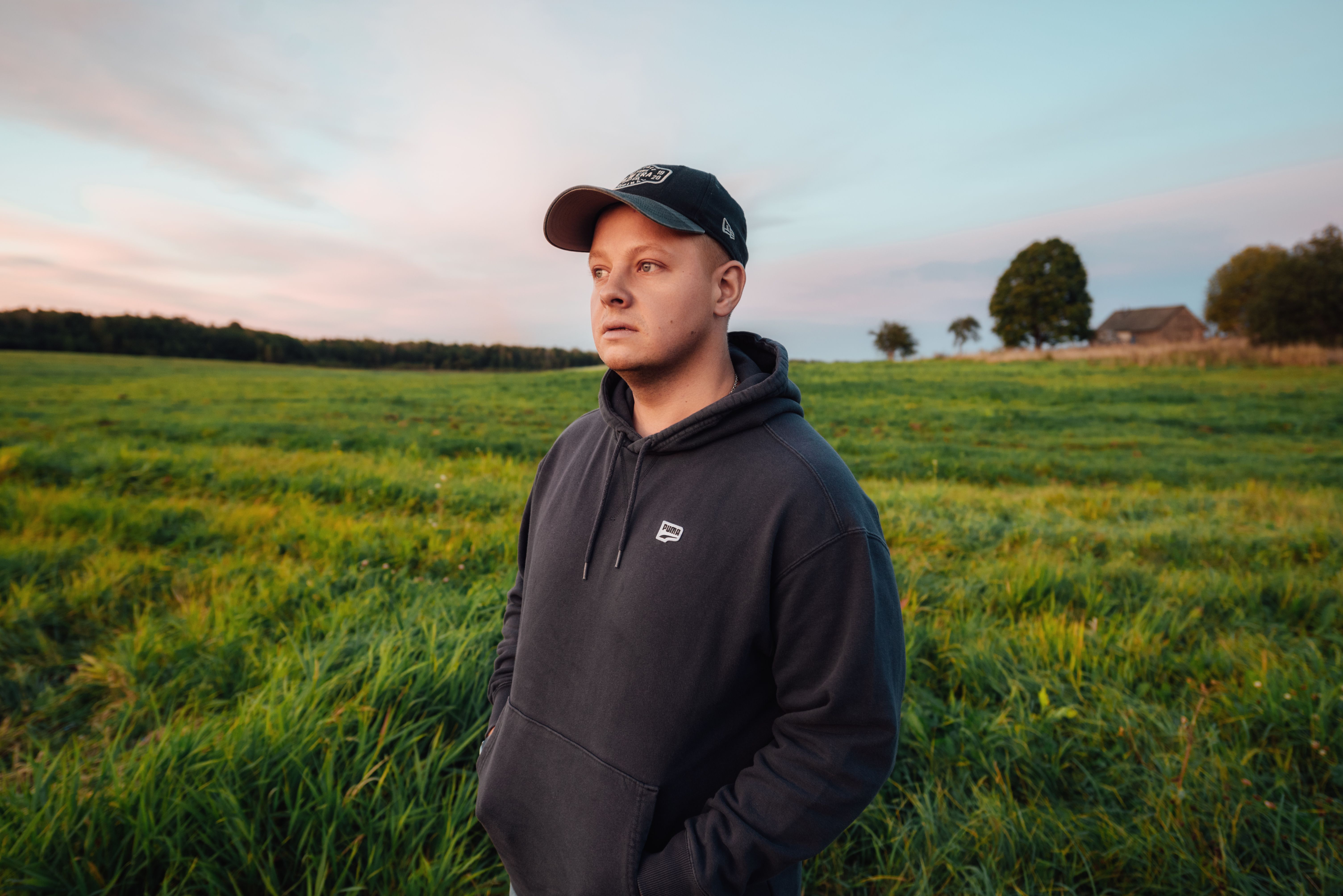 A man in a black hoodie and cap stands in a serene green field under a colorful sky, exuding tranquility.