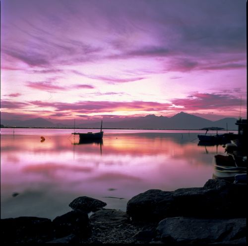 Free Beautiful sunset reflecting on a calm lake with silhouetted boats and distant mountains. Stock Photo