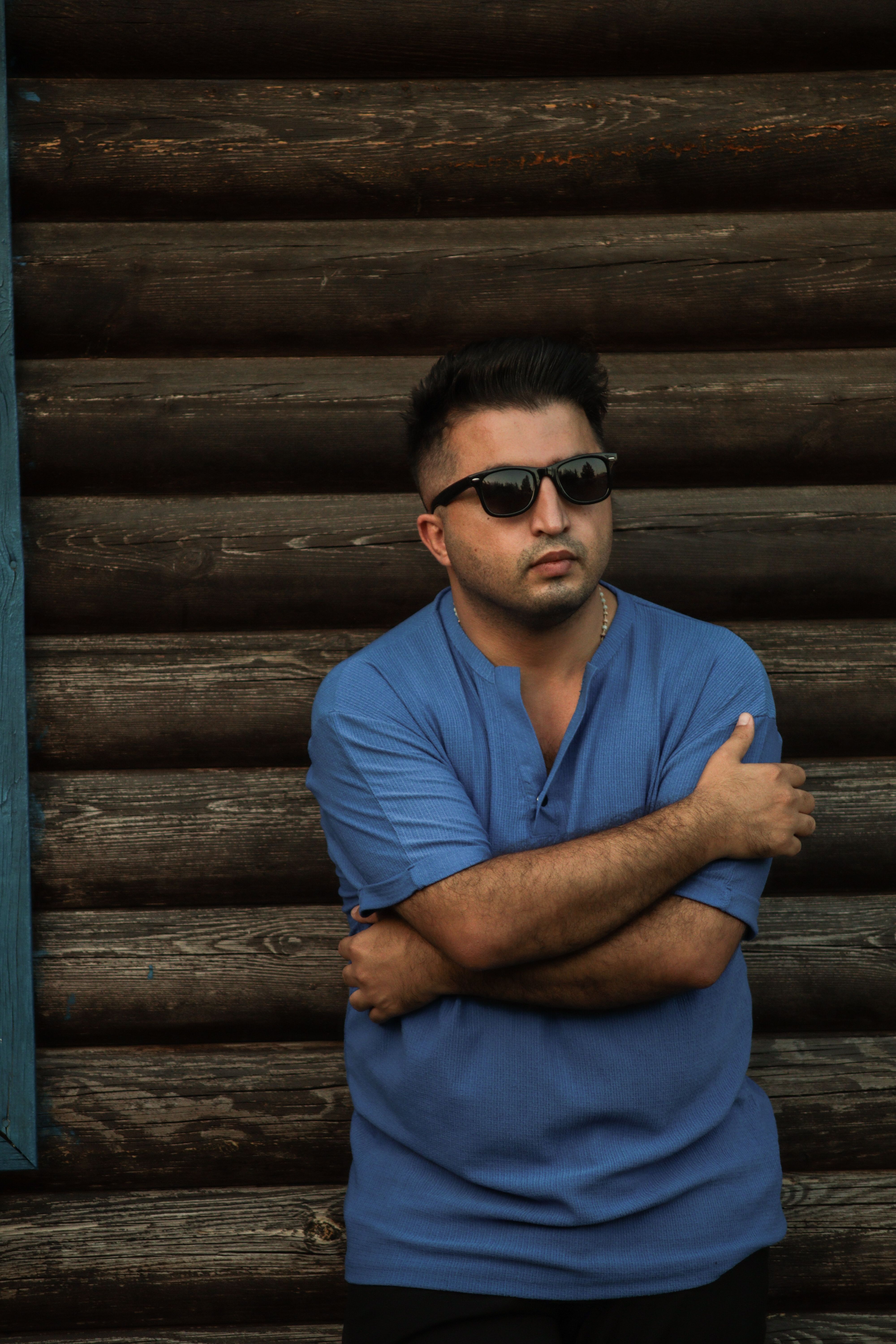 Man in blue shirt wearing sunglasses poses against a rustic wooden wall.