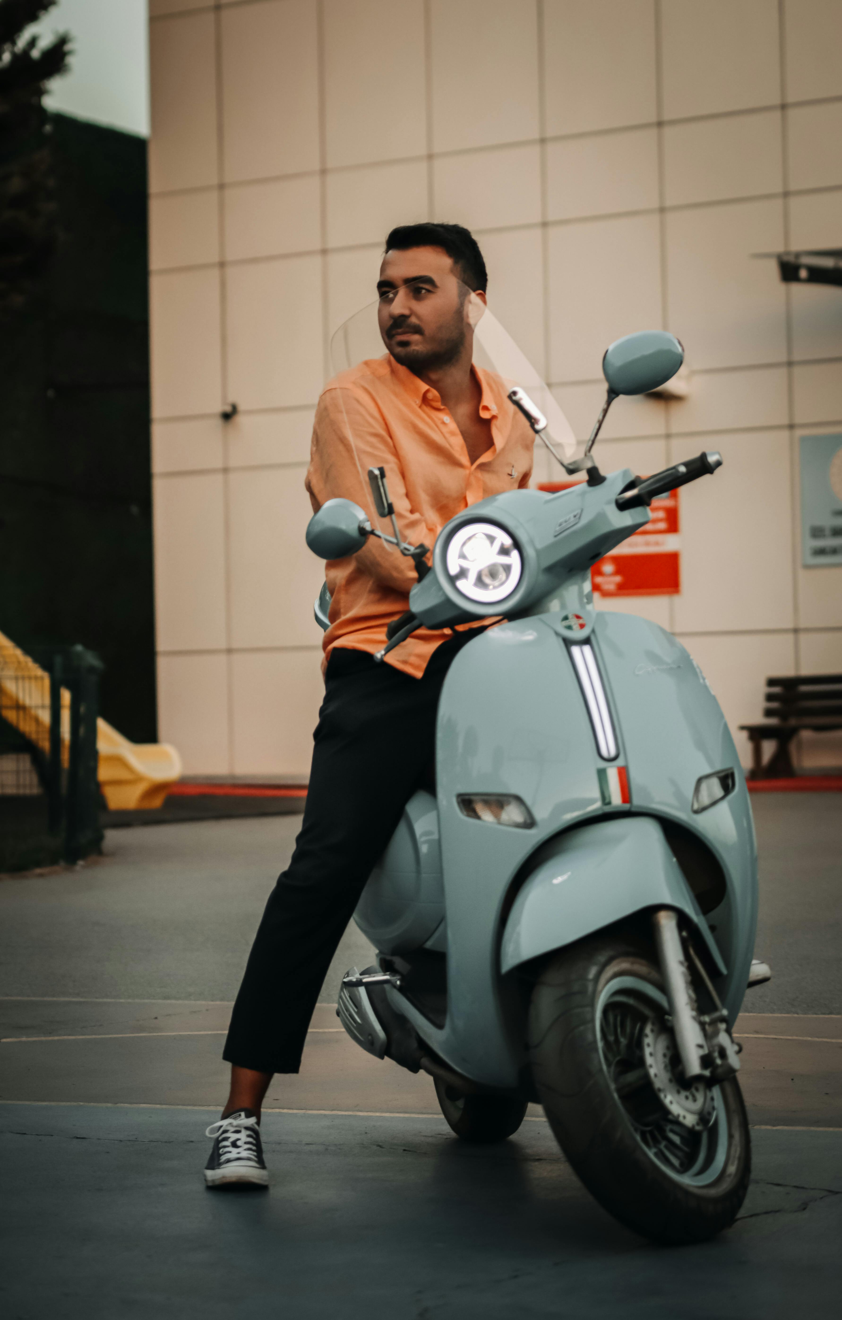 A young man leans on a retro-style scooter in an urban environment, exuding a relaxed and stylish vibe.