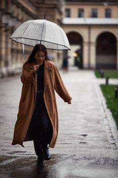 A woman in a brown coat walks under an umbrella on a rainy day in an urban setting.