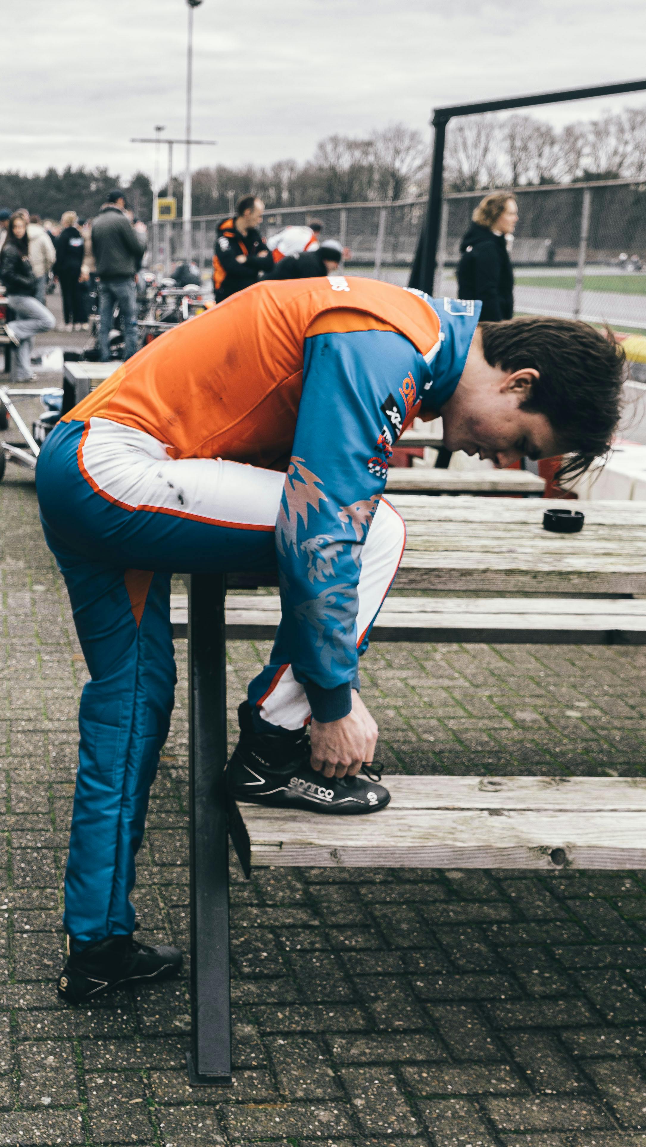 A young racer in colorful gear ties his shoe, preparing for karting at Berghem, Netherlands.