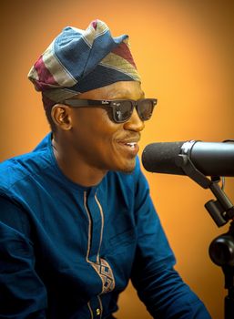 A man wearing traditional attire with sunglasses speaks into a microphone.