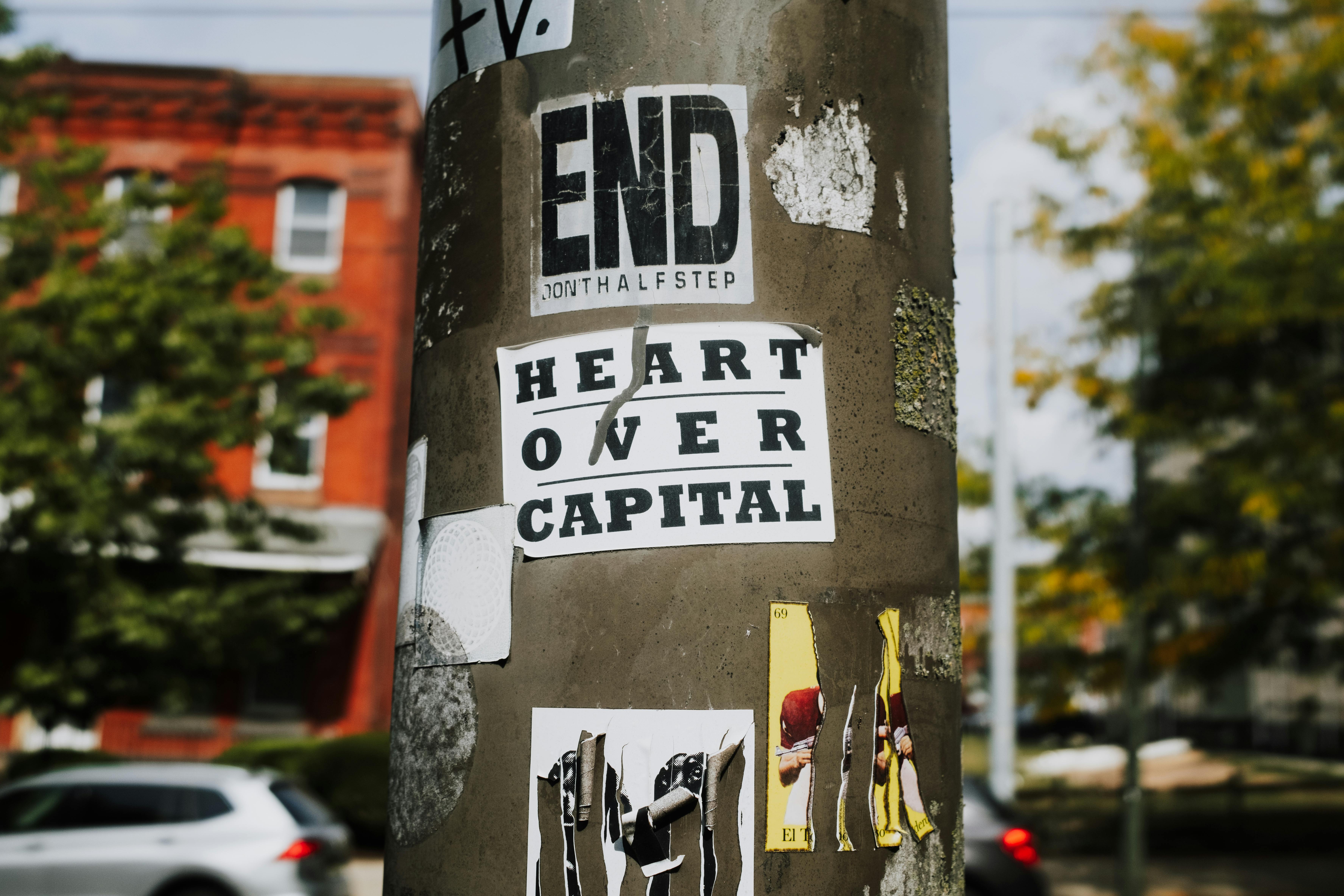 Street art and stickers on a utility pole in Philadelphia with the message 'Heart Over Capital'.