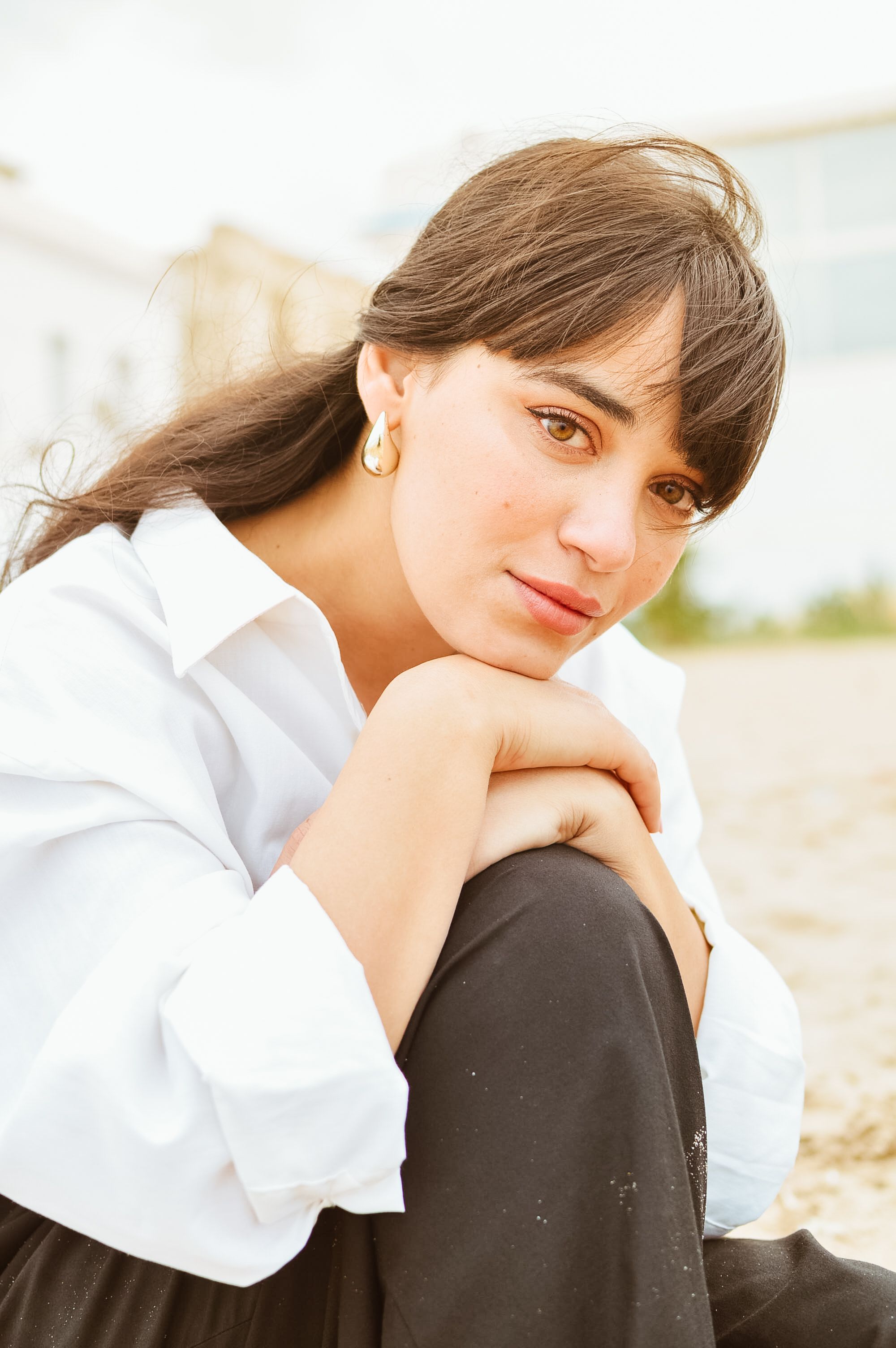 A woman in a white shirt, sitting on a beach, looking thoughtfully at the camera.