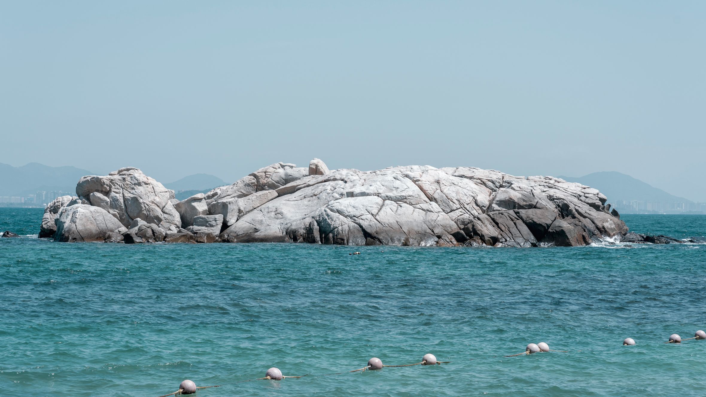 Tranquil scene of a rocky island surrounded by clear blue ocean water with distant mountains.
