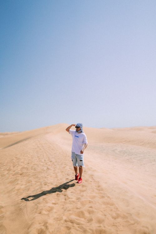 Free A man in casual attire walks across the sandy dunes of Dubai desert under a bright blue sky. Stock Photo