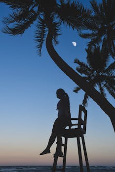 黄昏时分，多米尼加共和国的海滩上，一个女人坐在救生员椅上，剪影对着天空中的月亮和棕榈树