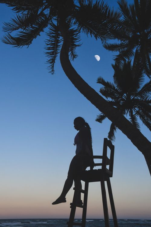 Free Silhouette of a woman sitting on a lifeguard chair against a twilight sky with moon and palm trees, Dominican Republic. Stock Photo