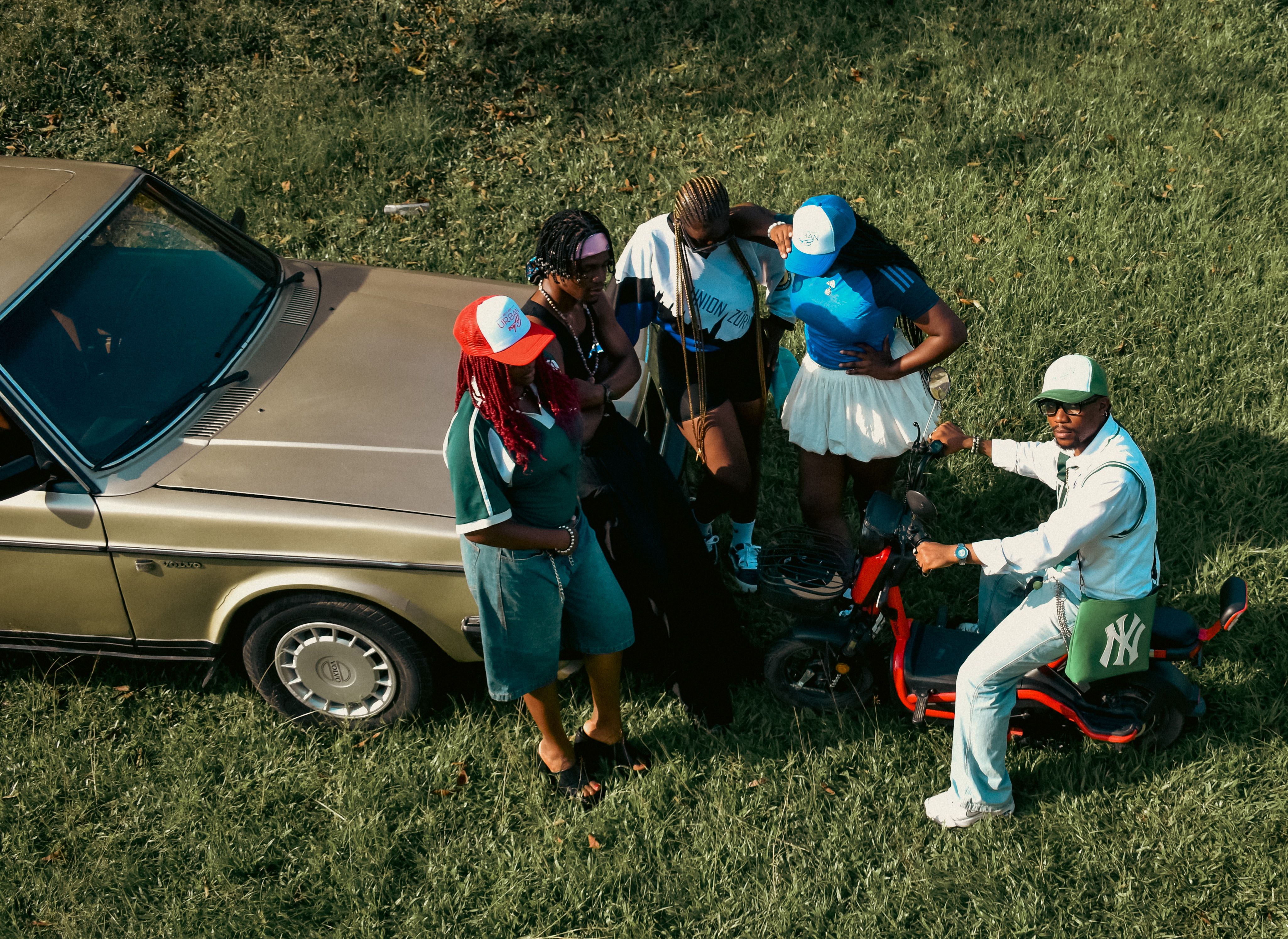 A group of friends gathers by a vintage car and scooter in a grassy outdoor setting.
