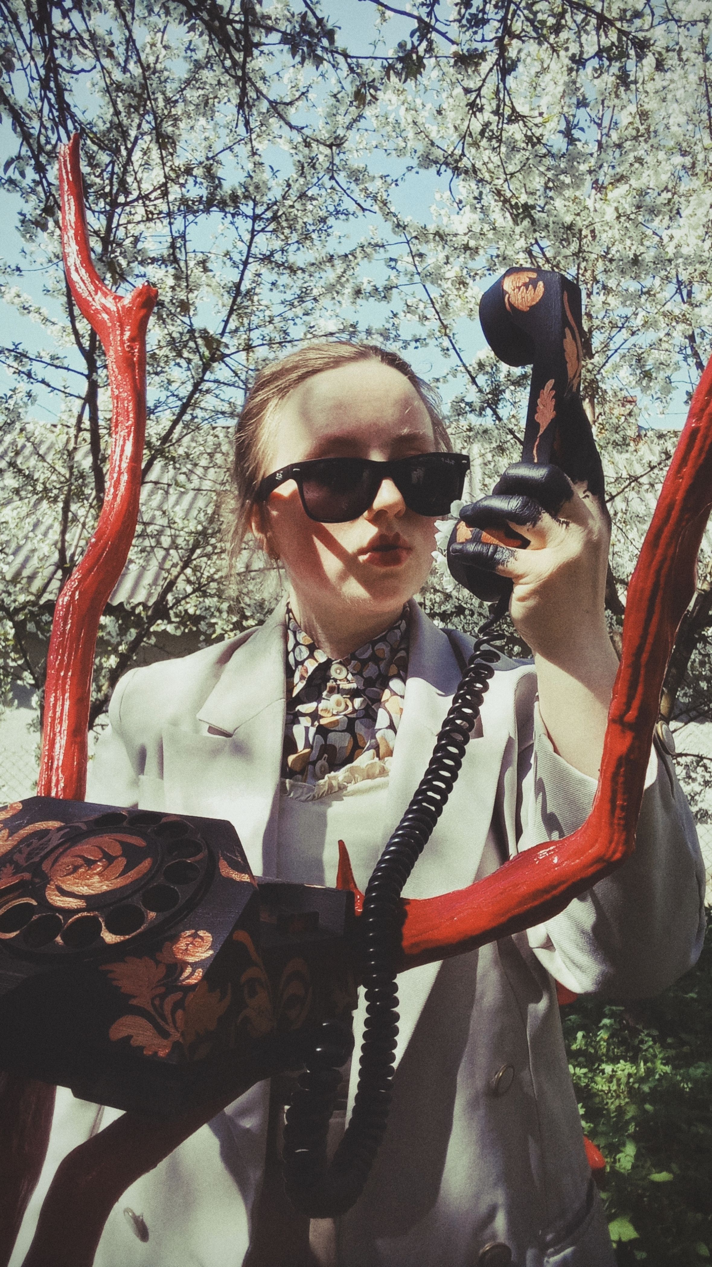 Stylish woman in sunglasses holds vintage phone outdoors with unique artistic installation.