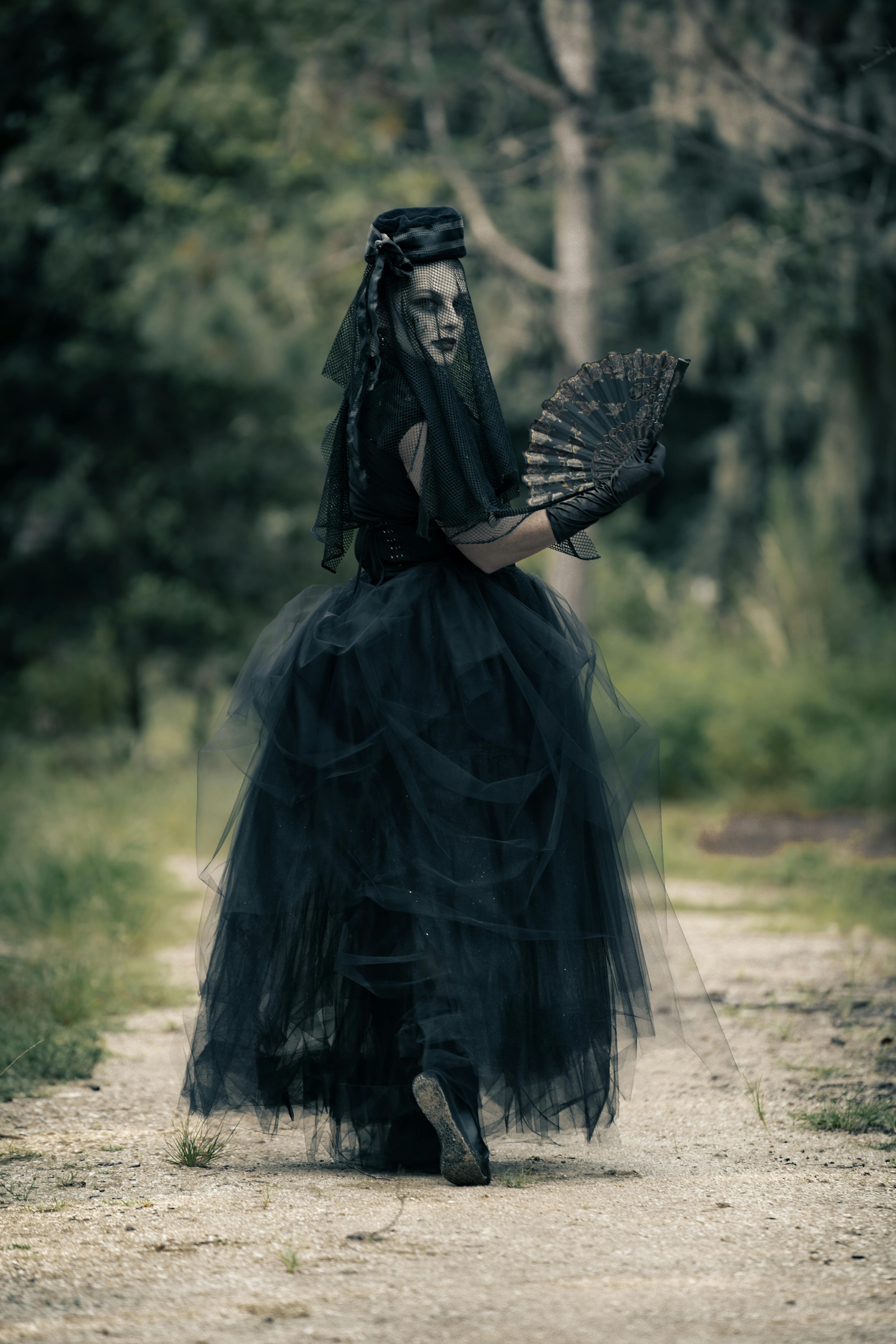Woman in a gothic black gown and veil holding a fan in a mysterious outdoor environment.