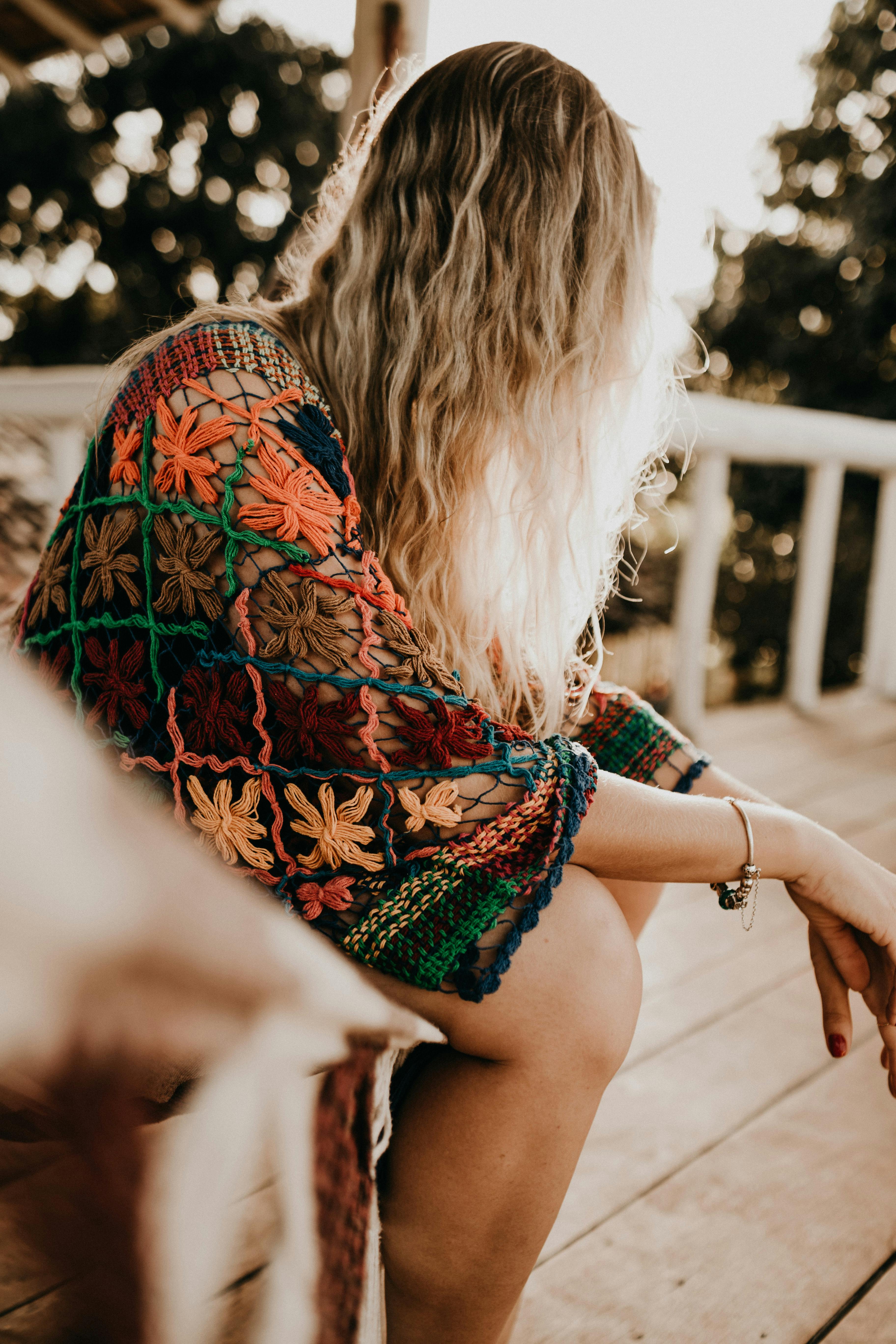 A woman with blonde hair sits on a wooden deck wearing a colorful knit shawl at sunset.