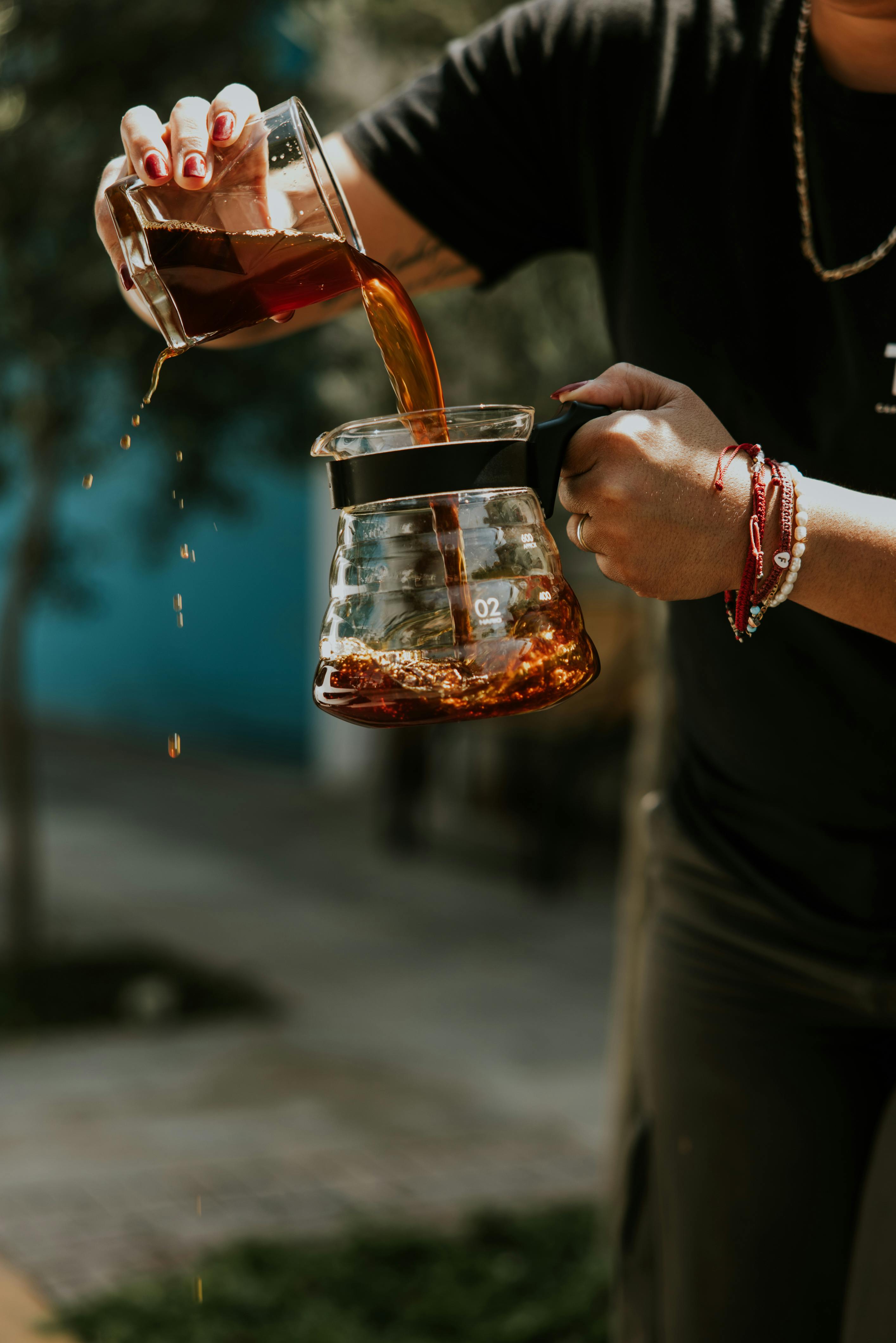 A person pouring fresh coffee into a glass jug in an outdoor setting with greenery.