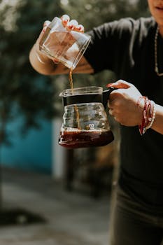 A person pours coffee from a small pitcher into a carafe outdoors, capturing the essence of coffee making.
