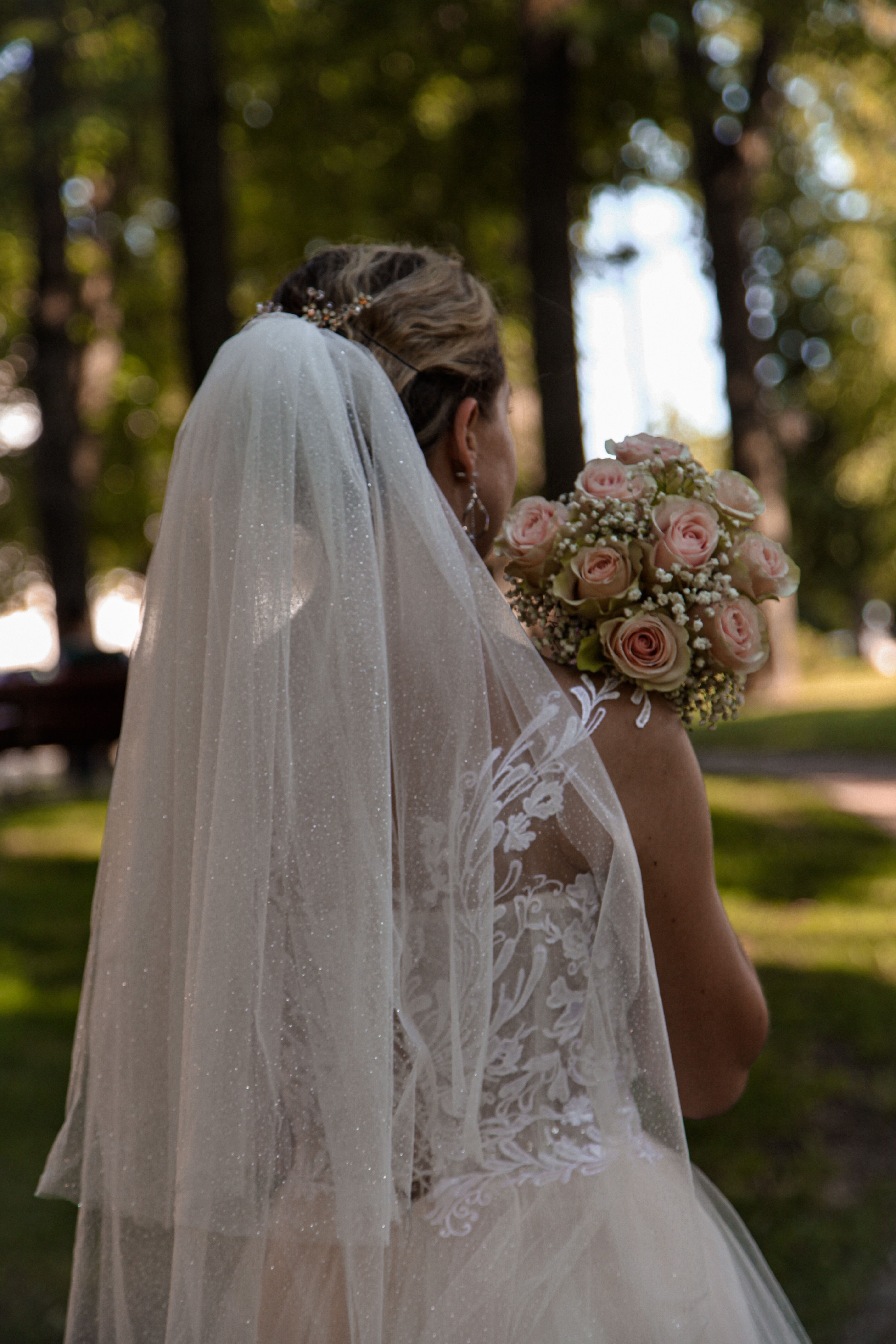 Bride in elegant dress holds bouquet in sunlit forest, captured from behind.