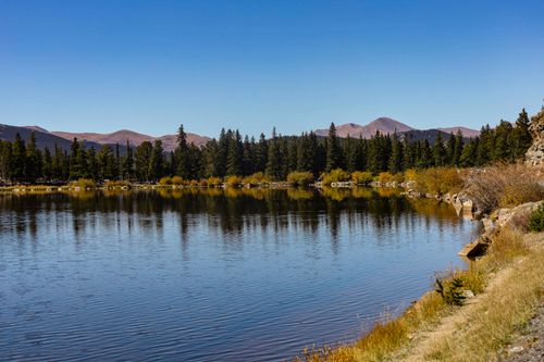 Free A peaceful mountain lake surrounded by forest and distant peaks under a clear blue sky. Stock Photo