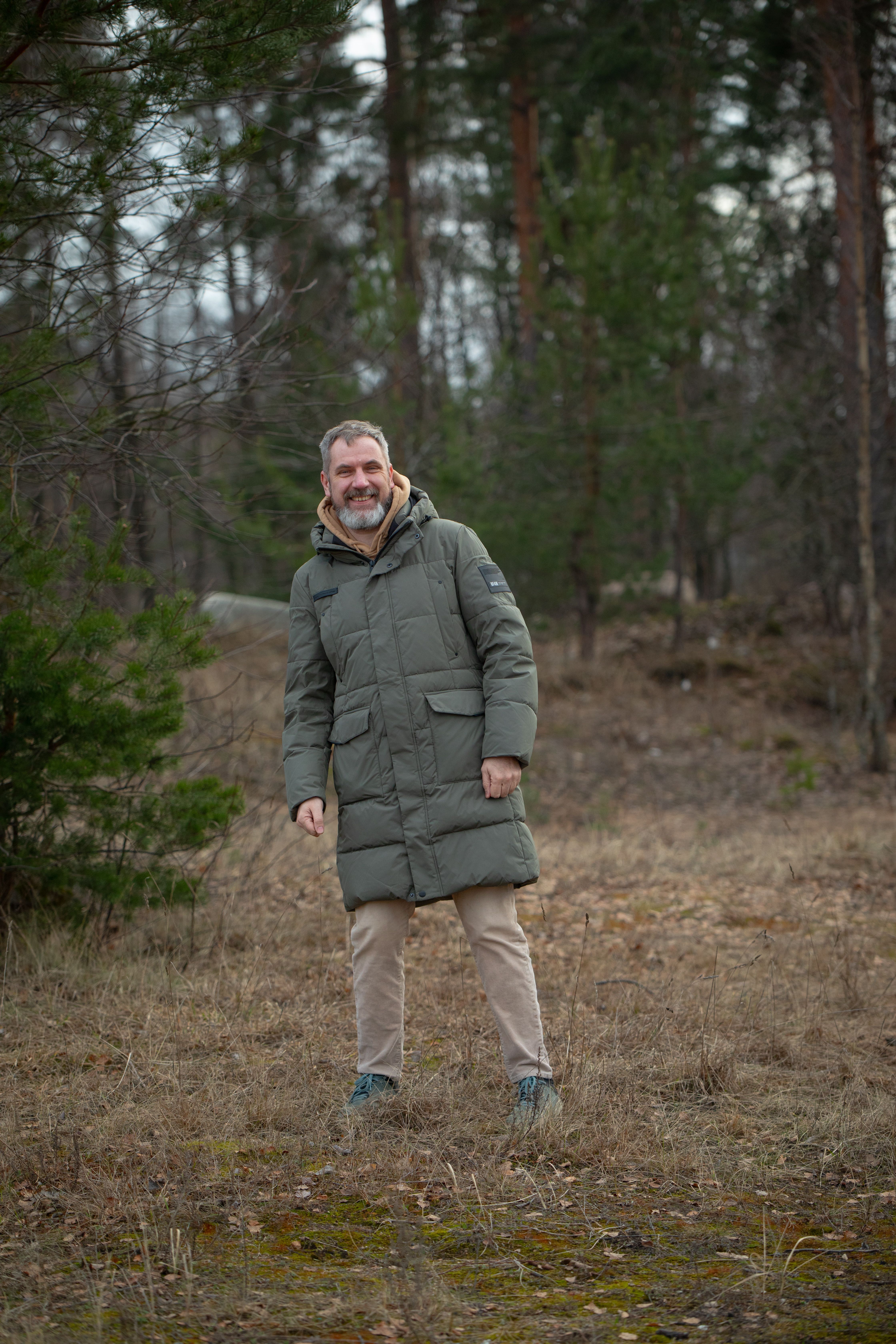 Adult man wearing a green winter coat in a forest setting, showing a relaxed and happy expression.