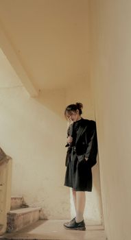 A young adult in a dark outfit standing thoughtfully in a dimly lit indoor stairwell.