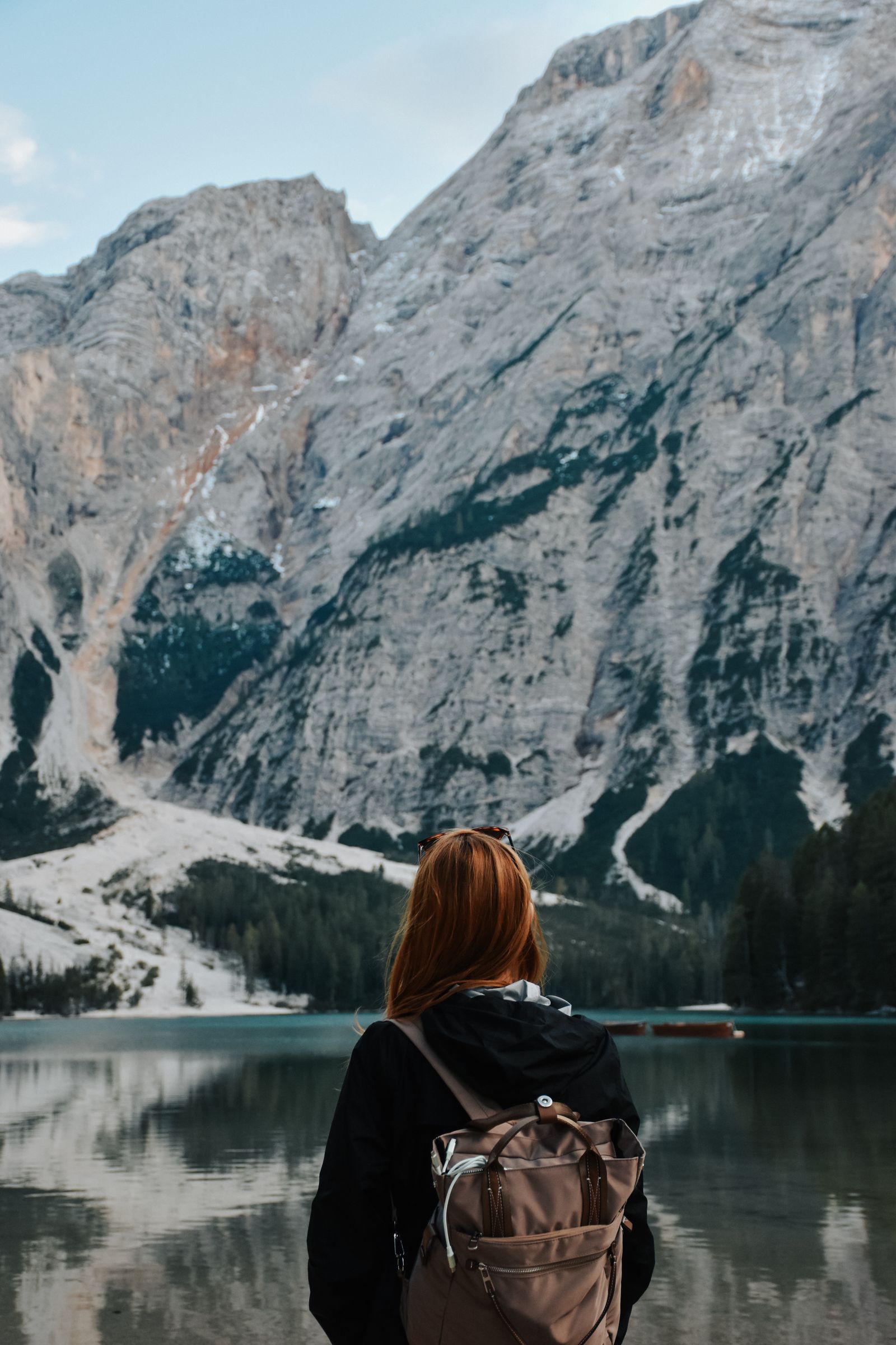 A woman with a backpack standing before a still mountain lake beneath towering peaks