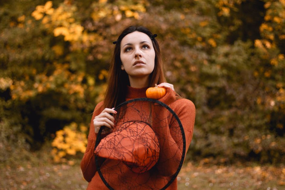 Adult woman holding a pumpkin and witch hat in a fall forest setting.