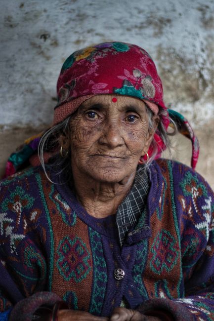 Elderly woman in a colorful headscarf and sweater, looking directly at the camera with a thoughtful expression.
