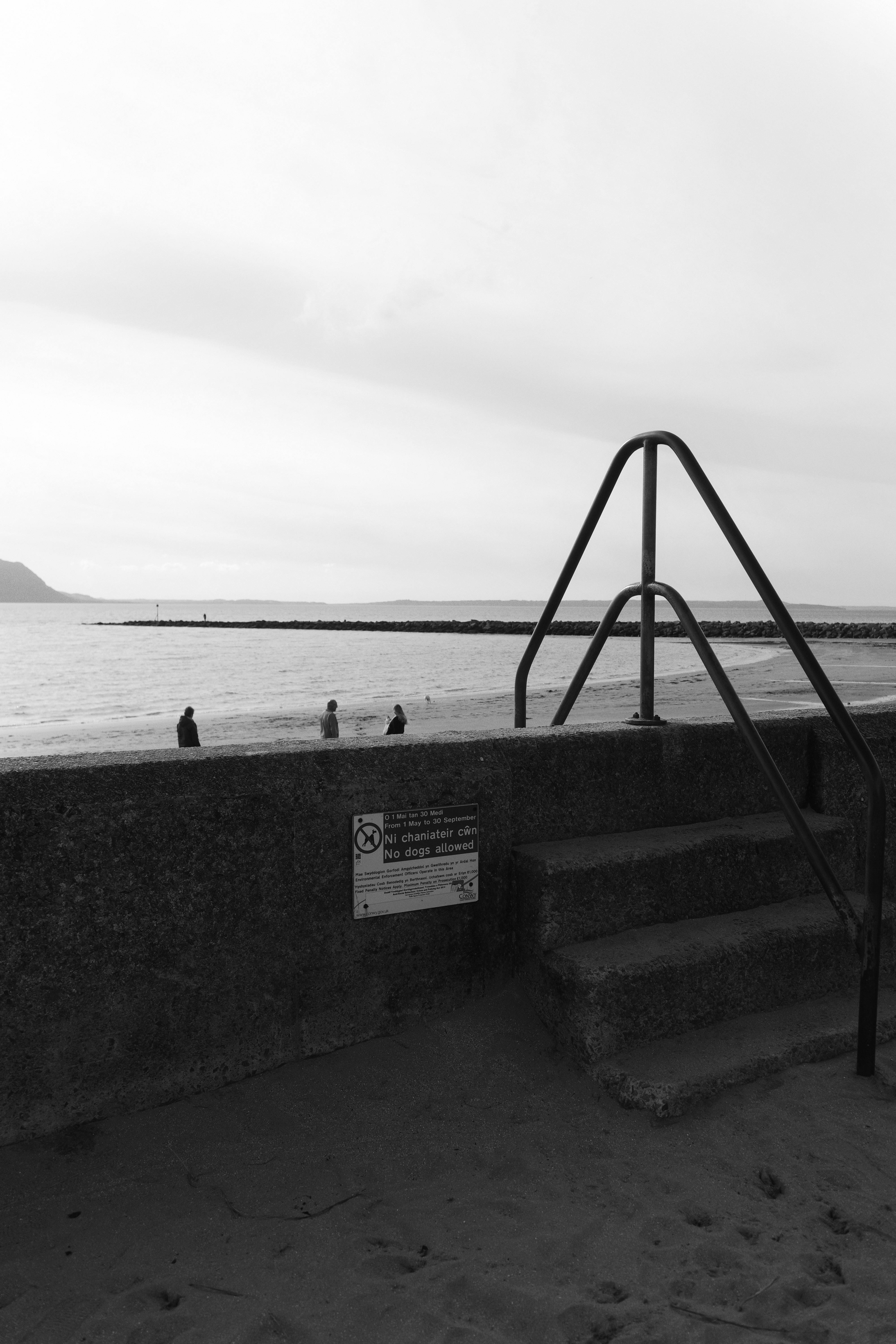 Monochrome beach view with people and stone staircase under cloudy sky.