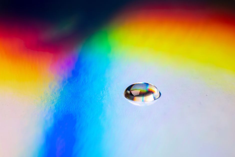 A colorful water droplet reflects prismatic rainbow hues on a smooth surface.